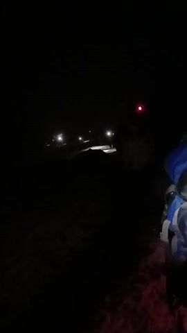 The video shows a group of people hiking in the dark. They are using headlamps to light their way. There is snow on the ground, and the person recording the video is walking behind them. The person recording has a blue backpack. One of the hikers has a red light on the back of their headlamp. The speaker states Kilimanjaro.