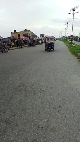 The video shows a street scene, possibly a marketplace, with several people and vehicles. The camera is oriented vertically, with the ground running horizontally across the screen. In the foreground, a person in a red top and dark pants walks away from the camera. Behind them, a blue and white tuk-tuk is parked, with various goods displayed on shelves and tables around it. Several market stalls with colorful awnings are visible, some with people gathered around them. A motorcycle is seen parked near one of the stalls. In the background, a large billboard with a picture of a man and text is displayed. The sky is overcast.