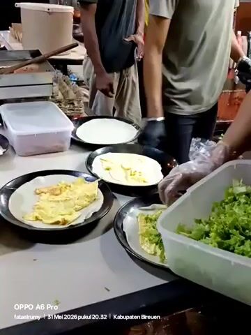 The video shows a man preparing food on a table. He is wearing gloves and adding lettuce to what appears to be eggs on top of what seems like wraps. The table has a white surface and is situated outdoors. There are several containers and plates visible on the table. Other people are in the background, moving around, but the focus is on the man assembling the food.