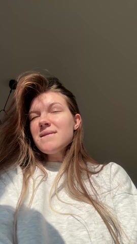 A young woman with long, light brown hair is standing in front of the camera, singing along to music and making faces. She is wearing a light grey sweater. Sunlight is shining on her face from the side. She sticks her tongue out, poses, and runs her fingers through her hair. A modern light fixture is visible on the ceiling behind her.