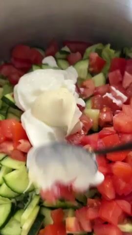 A close-up shot shows a salad with chopped tomatoes and cucumbers being mixed with a creamy dressing using a spoon. The salad is in a large silver pot or bowl, and the ingredients are vibrant red and green. The spoon is blurred as it moves through the salad, indicating rapid motion. The lighting is bright, highlighting the fresh ingredients.