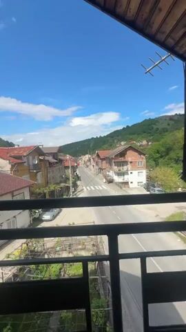 The video shows a street scene from a balcony, with a small town nestled between green hills. The sky is bright blue with scattered white clouds. The street is mostly empty, with a car parked near a crosswalk. Several buildings line the street, varying in color and construction. The balcony has a black metal railing.