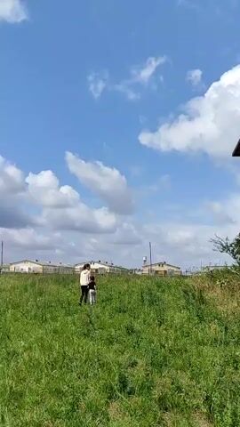 A wide shot shows a rustic, weathered wooden house with a sloping roof and multiple windows, some of which are broken. The house appears to be in disrepair. In front of the house, a grassy slope is visible, and two figures, one adult and one child, are walking across it. The sky is blue with scattered white clouds, and the overall environment suggests a rural or abandoned setting.