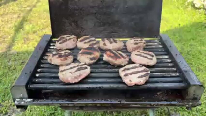 The video shows a grill with ten small, round patties cooking on it. The grill is old and black, with a hinged lid. The patties are arranged evenly across the grill and are marked with dark grill lines. Some are more browned than others. There is smoke rising from the grill. The grill is sitting on a stand in a grassy outdoor setting.