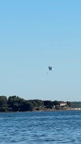 A person is parasailing over the ocean with a green tree-covered coastline in the background. The sky is clear and blue.
