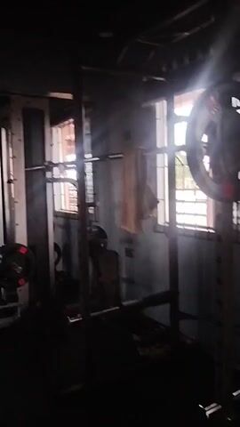 The video shows the interior of a gym. In the foreground, a weight rack is visible with weights on it. Further back, a large mural of a muscular figure is painted on the wall. Various gym equipment, including weight machines and benches, are scattered throughout the space. The lighting is dim, with natural light coming in from windows on the right side of the frame. The floor is covered with dark rubber mats. The camera is moving, panning across the gym.