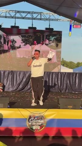 A man wearing sunglasses, a white shirt with a black logo, and black pants with red stripes is standing on a stage. He is holding a white object in his right hand and a microphone in his left. He appears to be singing or speaking to an audience. The stage is decorated with confetti and has a large screen behind it showing a video of the man on stage. There are Colombian flags on the front of the stage. The environment appears to be outdoors during the daytime, with a tent or canopy overhead.