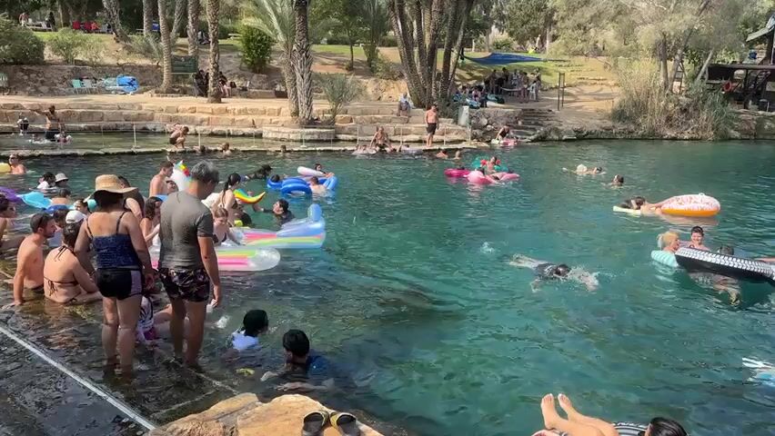 This video captures a busy outdoor swimming area. In the foreground, a group of people are gathered at the edge of a large, clear, turquoise pool. Some are standing in the shallow water, while others are sitting on the stone edge. A variety of inflatable pool floats, including a large unicorn float, are scattered throughout the water, with people relaxing on them.

The pool is surrounded by a tiered stone structure, and beyond that, lush greenery with numerous palm trees and other foliage. In the background, more people are visible relaxing on lounge chairs and under umbrellas in shaded areas. The overall atmosphere suggests a popular recreational spot on a warm, sunny day, with many individuals enjoying the water and the pleasant environment. The water appears to be shallow in the foreground and deepens further out. People of various ages are present, suggesting a family-friendly location.