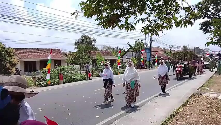 A street parade is taking place under a bright sky. Several people are walking in a procession along the right side of a paved road. They are dressed in traditional clothing, including long skirts and head coverings, and some are carrying small Indonesian flags. In the background, there are houses decorated with Indonesian flags and banners. A horse-drawn cart and a motorcycle are also part of the parade. Trees with green foliage line the top of the frame, obscuring some of the view of the sky and overhead wires. The overall atmosphere appears festive.