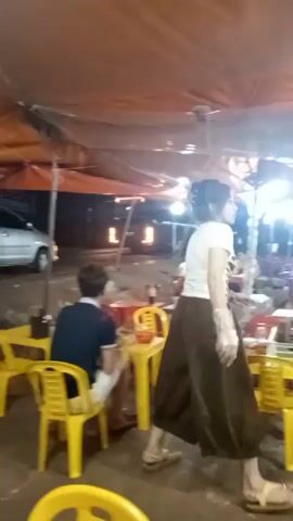 The video shows an outdoor food stall. There's a young woman wearing a white top and brown pants walking towards the stall. The stall is made of wood and has a glass display case containing various cooked foods, including what appears to be fried chicken and other meat dishes. Beside the stall is a stack of plates and some bowls. Several tables and chairs are set up around the stall, with some people seated. The area is covered by a large tent, and there are lights illuminating the space.
