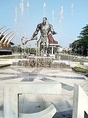 The video shows a bronze statue of a muscular man, possibly representing a prehistoric human, holding a large club. The statue stands in the center of a circular fountain, with water spraying upwards around it. In the background, there is a structure with large, tusk-like elements. The scene is bright and sunny, suggesting it is daytime. The surrounding area appears to be a park or public space with roads and trees. There are white concrete benches in the foreground.