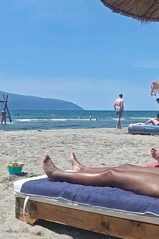The video shows a beach scene on a sunny day. A person is lying on a beach chair with blue cushions. Their bare legs are visible. In the background, there are other people on the beach, some sunbathing on beach chairs, others walking or swimming in the ocean. A wooden swing set is visible in the distance, partially submerged in water. There is also a child's bucket on the sand nearby. The sky is clear and blue.  A straw beach umbrella is partly visible overhead.