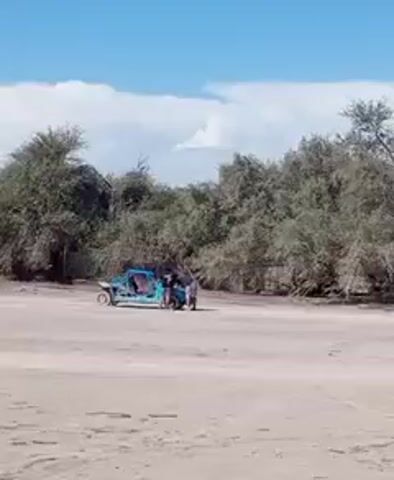 The video shows a group of people in a sandy, possibly beach-like environment with scrubby trees in the background under a partly cloudy sky. A blue off-road vehicle is parked on the sand, and several individuals are standing nearby, appearing to be working on or inspecting it. One person is mounting and starting a dirt bike, then rides it away from the group and the blue vehicle.