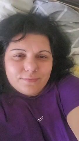 A woman with dark, curly hair and brown eyes is lying down and looking at the camera. She is wearing a purple t-shirt with a small beaded design on the left side of her chest. The background appears to be a bed with patterned sheets. The lighting is bright and comes from above. The woman is smiling slightly and her expression is relaxed. The video is a selfie, as evidenced by the woman holding the camera or phone herself.