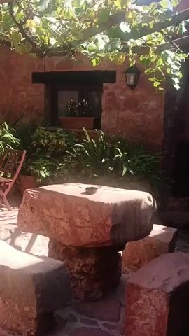 This video shows an outdoor patio area with a stone table and chairs set under a grape arbor. The patio is paved with brick and surrounded by a stone wall. There are several potted plants and a red chair visible. The video pans across the patio, showing different angles of the scene. The lighting suggests it is a sunny day.