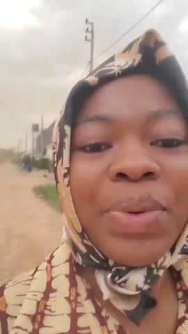 A woman with dark skin and brown eyes is looking directly at the camera, smiling, and talking. She is wearing a tan and brown patterned shirt and a tan, brown, and black patterned headscarf. The background appears to be a street with buildings and vegetation visible. The sky is overcast. The woman is likely recording a video selfie.