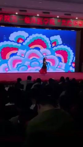 The video shows a person performing on a stage in front of an audience. The stage is covered in a red material. The performer is wearing a red dress and some sort of head covering. Behind the performer is a large screen with a colorful abstract design displayed. The audience is seated in rows and appears to be watching the performance. There is a sign above the screen with text in Chinese.