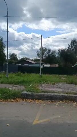 The video shows a rural landscape with a dirt road leading into a village. The road is bordered by green grass and trees. There are houses and fences visible, as well as a street lamp. The sky is cloudy. The video appears to have been filmed from a moving vehicle. The only visible sign of human activity is a red house with white trim, and a greenhouse with "ЦЕНТР" written on it, suggesting it may be a commercial establishment. There are no people visible in the video.