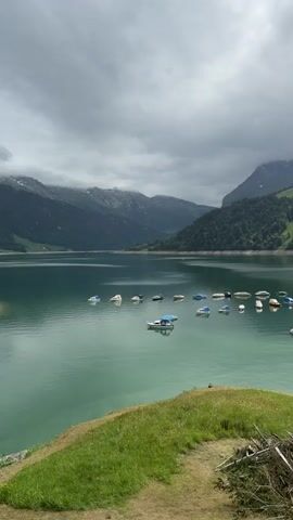 A vertical video shows an overcast mountainous landscape featuring a turquoise-green lake with numerous small boats, many covered, anchored across its surface. The camera slowly pans right, revealing a grassy foreground with a dirt path and a pile of cut branches. The lakeshore, with some exposed drier ground, becomes visible, including a small yellow structure and a dock with a few more boats near it on the right side of the frame. Lush green, forested hills and mountains rise from the lake under a cloudy sky.