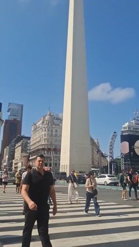 The video shows a daytime shot of the Obelisco de Buenos Aires, a tall, white obelisk monument located in Buenos Aires, Argentina. Several people are walking across a crosswalk in the foreground, in front of the obelisk. Cars and taxis drive by on the surrounding streets. In the background are buildings of various heights and architectural styles, as well as digital billboards. The sky is blue with some scattered clouds. The atmosphere appears sunny and bustling.