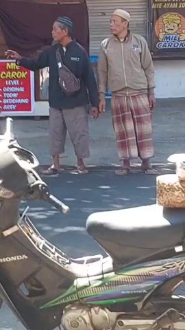 A street scene unfolds. In the foreground, a black Honda motorcycle is parked facing left, its front wheel visible. Behind the motorcycle, three men stand on a paved street in front of a building with a red banner. The man on the left wears a patterned shirt and a sarong. The man in the center, facing right, wears a dark jacket, grey pants, and a satchel across his chest. He appears to be gesturing with his left hand. The man on the right, facing left, wears a light-colored jacket and a sarong. The red banner above them reads "BAKSO GEMOY" in large yellow letters, with smaller text below that is partially obscured but includes "MIE CAROK." A motorcycle rider is blurred in motion to the right of the central man, moving away from the camera. The environment appears to be outdoors with sunlight casting shadows.