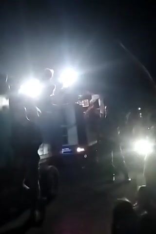 The video shows a group of people at an outdoor event at night. Bright lights illuminate the scene. There is a truck with people on top of it. Several people are dancing, including one person in a red dress and another in a blue dress. There are also people standing and watching. The environment appears to be a street or an open area.