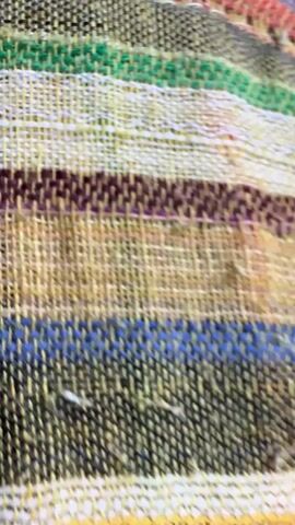 The video begins with a close-up, angled shot of a textured, woven fabric with horizontal stripes of various colors including yellow, green, maroon, blue, and white. The camera then pans to the right, showing more of the fabric. The scene then shifts to an interior view of a crowded train carriage. In the foreground, a young woman with blonde hair and tattoos on her chest is seen from the chest up, wearing a white floral print dress and a white bracelet on her left wrist. She is looking to her left, out of frame. The focus then shifts to the full interior of the train carriage, showing rows of seats occupied by numerous passengers. The lighting appears to be natural, coming from the windows and the overhead lights. The overall atmosphere suggests a busy public transportation environment.