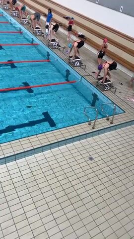 A swimming race begins. Several swimmers wearing swim caps and racing suits are positioned on starting blocks at the edge of an indoor swimming pool. They are lined up along different lanes, marked by floating lane dividers. A coach stands nearby. At the start signal, the swimmers dive into the water and begin to swim. The camera angle is elevated, providing a clear view of the pool and the swimmers as they enter the water. The pool is surrounded by tiled flooring.