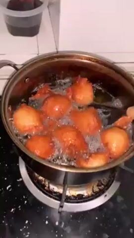 The video shows a pan on a stove filled with bubbling oil and several golden-brown, round pastries frying inside.  A person uses black tongs to turn the pastries in the oil. The stove is black with some white staining.  A plastic container with a dark liquid is visible in the upper left corner of the frame.