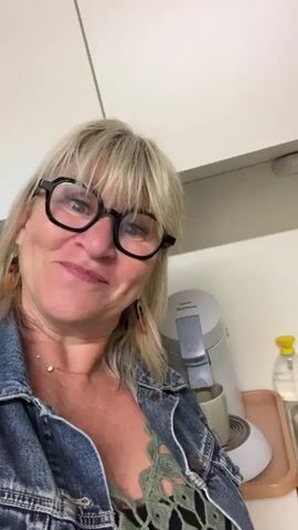 A blonde woman with shoulder-length hair and bangs smiles warmly into the camera, taking a close-up selfie-style video. She wears black-framed glasses, a denim jacket, and a green, possibly lacy or crocheted, top. A delicate gold chain necklace with a small pendant and a patterned earring are visible.

Behind her, in a kitchen setting, a light grey Philips Senseo coffee machine is actively dispensing coffee into a white mug placed on a small pinkish-orange tray. To the right, a stainless steel sink is partially visible, next to two dispenser bottles: one with a yellow pump top and another larger clear bottle with an illegible label. The background consists of clean white kitchen cabinets or wall panels.

The woman appears happy and content, seemingly enjoying a casual moment, possibly a coffee break or morning routine. The scene is located in La Fouillouse, France.