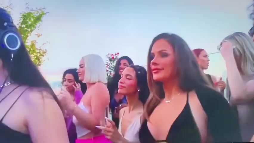 The video shows a group of young women at an outdoor gathering. The women are casually dressed, many in revealing clothing, and are holding drinks. The environment appears to be a rooftop with city buildings visible in the background, suggesting a party atmosphere. Some of the women are talking and interacting with each other. The overall vibe is celebratory and social.