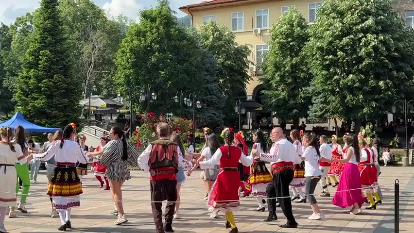 A large group of approximately thirty to forty people of various ages are holding hands and dancing in a large circle on a paved outdoor square. Many participants, particularly women, are wearing traditional folk costumes with embroidered blouses and colorful skirts, while others are in casual modern clothing like shorts, t-shirts, and dresses. The dancers move in a counter-clockwise direction, performing a synchronized folk dance with steps and arm movements. In the background, there are green trees, a light-colored building, and some small businesses with umbrellas. A blue tent is visible on the far left side of the square. The scene is brightly lit by natural daylight.