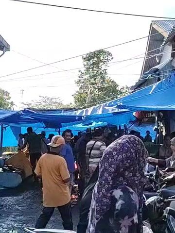 The video shows an outdoor market with people milling about and vendors standing behind tables. Blue tarps provide cover over the vendors and shoppers. Several motorcycles are parked nearby.  People are mostly interacting with vendors at their stalls. The market area appears wet. The light is bright, suggesting it is daytime.