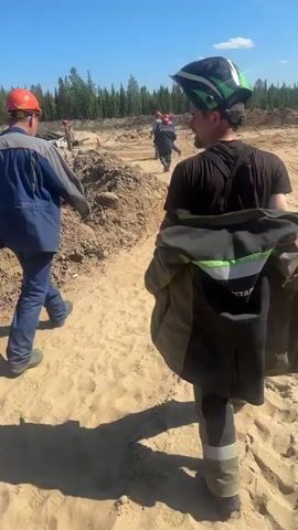The video shows a group of construction workers in a sandy, outdoor environment. The sun is shining brightly, casting distinct shadows on the ground. In the foreground, a person wearing a welding helmet and a dark jacket with reflective green stripes is walking away from the camera, carrying their jacket over their shoulder. To the left, another worker wearing an orange hard hat and a blue and gray uniform is walking towards the left side of the frame. Further in the background, several other workers, also wearing hard hats, are visible, some appearing to be engaged in work or walking in the distance. The terrain is uneven and appears to be a construction site with piles of dirt and sand. In the far background, a line of trees is visible under a clear blue sky, suggesting a rural or undeveloped area. There are some blue structures and possibly equipment scattered across the background. The overall impression is a work-in-progress environment under good weather conditions.