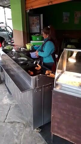 A woman in a blue shirt and black apron is preparing food at an outdoor food stand. She is using tongs to take something from a blue bowl and place it into a deep fryer containing chicken pieces. There is a warming case with french fries and other food next to the fryer. The food stand is next to a building with green walls and a rolled-up shutter. A car is partially visible in the background. A menu board is behind the warming case. Another person wearing a winter hat can be seen in the background, preparing food on a counter. The ground is paved with stone tiles.