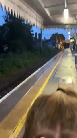 The video shows a train platform at dusk. The platform is covered by a white awning, and the yellow line marking the edge of the platform is visible. Train tracks run along the side of the platform, with greenery visible beyond the tracks. The sky is a dark blue, and the scene is illuminated by overhead lights. A person's back is visible in the foreground.