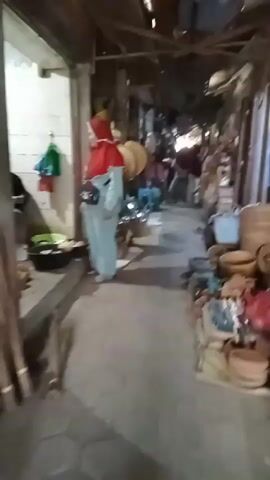 This is a video taken from a slightly elevated perspective, looking down a narrow, covered market alley. The ground is dirt or concrete. On either side of the alley are stalls displaying various goods. The stall on the left shows baskets, what appear to be metal bowls or pots, and some greenery. A person wearing a light blue garment and a red head covering is visible on the left, facing away from the camera and looking into the market. The stall on the right displays many woven baskets of different sizes and shapes, as well as some earthenware pots and other items. Further down the alley, several people are visible, some of whom appear to be interacting with each other. They are dressed in traditional clothing. The ceiling of the market appears to be made of corrugated metal or wood. The lighting is dim, with some brighter areas further down the alley, suggesting natural light sources. The overall impression is that of a busy, traditional market.