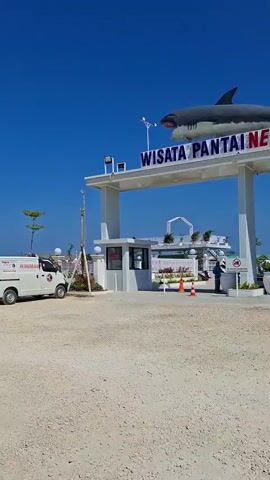 A white van is parked on the left side of the frame, with the text "SIAGA" visible on its side. A large shark sculpture is mounted on an archway that reads "WISATA PANTAI NEW HARYON". A person stands at a kiosk under the archway. To the right, several motorcycles are parked. Beyond the archway, there is a white fence with decorative elements, and beyond that, a white building structure. A silver car is parked in the middle ground, with its doors open and a person visible inside. The ground is covered with gravel and sand, and the sky is clear blue.