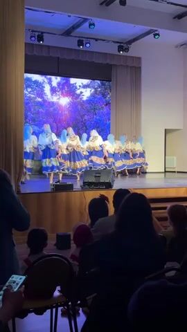 A group of approximately ten young girls in matching folk costumes performs a dance on a stage in a theater or auditorium. The costumes consist of a light-colored blouse, a blue and floral patterned skirt, and a light blue headscarf. The stage backdrop features a large screen displaying an image of trees with sunlight shining through. The dancers are performing lively steps, including jumps and spins. An audience, mostly silhouetted, watches from below. A person in the front row is using a smartphone, likely recording the performance. Stage lighting is visible above, casting a blue hue on the scene. A pair of speakers is positioned on the stage in front of the backdrop.