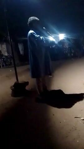 A person in a blue robe is standing in the middle of a dirt area at night, playing a stringed instrument under the illumination of a bright light. The person's shadow is cast on the ground. A pole stands next to them, with a dark object at its base. In the background, there are blurred shapes, possibly of people and objects.