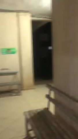 The video begins in a room with tiled floors and benches. There is a dark opening that may lead to another room. The video then pans to a glass surface, perhaps a window, showing a room with a large shelf containing various items. The camera moves quickly, showing another room lit by bright lights. The video ends with a shot of a blank tan wall. No people are visible in the video.