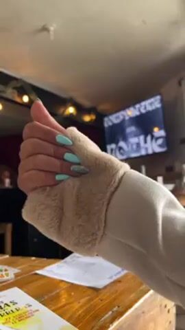 The video shows a woman's hand on a wooden table. The woman is wearing a light-colored sweater with a faux fur cuff. Her fingernails are painted a light blue color, except for one which has a glitter design. The hand is initially posed with the thumb up, and then the fingers are spread. On the table is a book with text in Spanish and yellow and green watercolor accents. Behind the hand, the background includes a television with a broadcast displaying the text "WHOA!" There are some string lights and other bar-type decor hanging in the background. The scene is dimly lit.