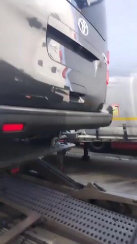 A black Toyota van is parked on a car transporter trailer. The camera is positioned low, looking up at the rear of the van. The van's tailgate is closed, and the taillights are visible. Underneath the van, a spare tire is mounted. The car transporter has a metal platform with a perforated surface. A large, white truck trailer is parked next to the transporter, partially visible in the background. The environment appears to be an outdoor loading or unloading area, possibly a car dealership or transportation hub. The lighting is bright, suggesting daylight.