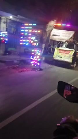 The video captures a nighttime street scene, likely in Southeast Asia, from the perspective of a moving vehicle, possibly a motorcycle. The street is paved with white lane markings visible. On the right side of the road, there are several food stalls or small shops illuminated by neon lights and white light boxes. One stall has a yellow banner with Indonesian text and a picture of food. Another stall displays a durian fruit. There are chairs set up outside some stalls, and a motorcycle is parked on the side. On the left side, there are buildings and a dark fence. The lighting is dim, and the movement of the camera creates a slightly blurred effect, but the general environment is clear.