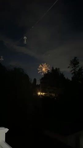 The video captures a nighttime scene with fireworks. The first frame shows a dark sky with some clouds. A thin cable with an object hanging from it is visible in the upper left. Fireworks are exploding in the distance, creating a bright, starburst effect. The second and third frames show more of the fireworks, with multiple explosions creating a dense cluster of lights against the dark sky. Trees and houses are silhouetted in the foreground. The fourth frame shows a single firework exploding, with bright yellow and orange sparks radiating outwards. The final frame shows a large, complex firework explosion with numerous bright sparks. The overall scene is dark and atmospheric, with the fireworks providing the only significant source of light.
