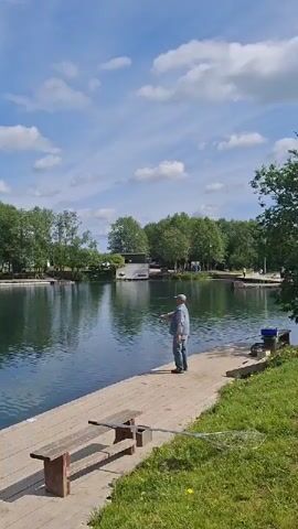 The video captures a tranquil outdoor scene at a lake in Kipen, Russia, on a bright, sunny day with a clear blue sky dotted with white, fluffy clouds.

The central subject is a man, dressed in a blue long-sleeved shirt and dark trousers, standing on a wooden dock or platform along the edge of the water. He is actively engaged in fishing, holding a fishing rod and casting his line into the calm, dark green water of the lake. Beside him on the dock, a wooden bench is visible, and further to his right, a blue bucket and a fishing net are placed, indicating his fishing gear.

The lake is bordered by a dense line of green trees, reflecting their foliage and the sky onto the water's surface. In the background, across the lake, a white building or structure is partially visible amidst the trees. The overall ambiance suggests a peaceful and relaxing leisure activity in nature. There is no visible text in the video.