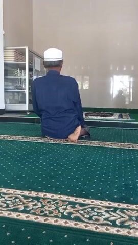 A person wearing a white kufi and a dark blue robe is praying on a green carpet inside a room. The person is shown from behind, kneeling and then bowing down, touching their forehead to the carpet. The room has a white wall and a glass cabinet filled with books on the left. Another prayer mat is visible in the background. The lighting appears to be natural light coming from windows.