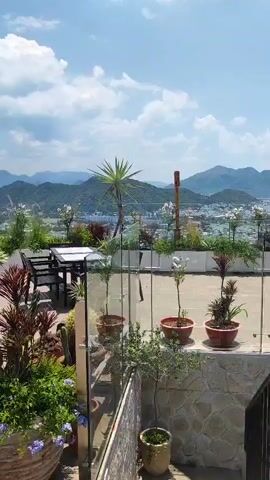 The video shows an outdoor rooftop seating area with a panoramic view of a city and mountains under a clear blue sky with scattered clouds. The area is furnished with tables and chairs, including high-top tables with bar stools and lower tables with chairs. Several potted plants and planters are strategically placed throughout the seating area, adding greenery and visual interest. In the foreground, there is a large potted plant with red and green foliage. The camera pans across the scene, showcasing the different seating arrangements and the expansive view. The lighting suggests it is daytime, likely sunny. No people are visible in the video.