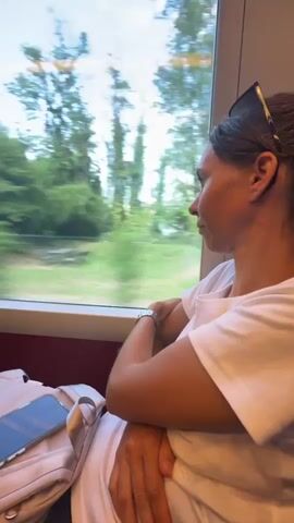 A woman with brown hair pulled back is sitting on a train, looking out the window. She is wearing a white t-shirt and a watch on her left wrist. Her arms are crossed. Next to her, on the seat, is a light pink backpack and a smartphone. The train is moving, as indicated by the blurred scenery outside the window. The scenery consists of green trees and a body of water with mountains in the distance under a blue sky with clouds. The interior of the train car is visible in the foreground with red seating.