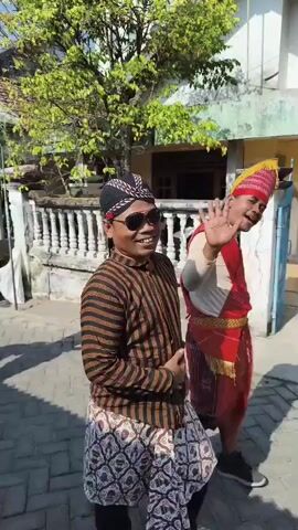 The video shows a group of men walking down a paved path in what appears to be a village. The men are dressed in traditional clothing, some in black with patterned sarongs and headwear, others in white with similar attire, and one man in a red and white striped shirt and black pants, carrying a whip-like object. They are walking in a procession, with some of them smiling and interacting with each other. The environment includes houses with tiled roofs, green trees, and a white fence. The sky is clear and blue, suggesting it is a sunny day. The overall atmosphere seems festive.